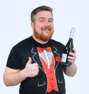 Brian Anderson wearing a tuxedo tee-shirt while holding a bottle of champagne and giving a thumbs up gesture.